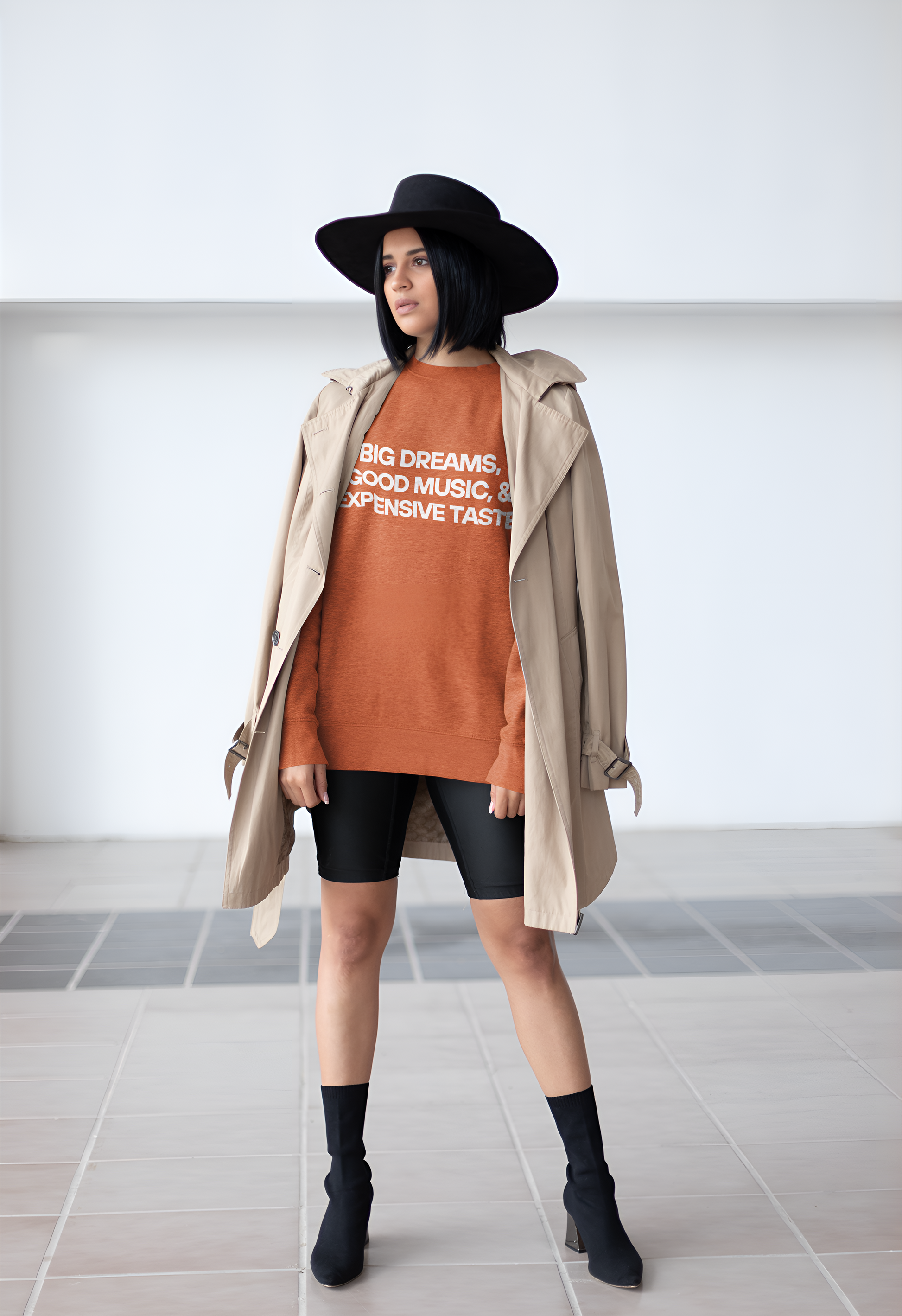 An Indian woman wearing a coral sweatshirt with "Big Dreams, Good Music, & Expensive Taste" written on it, a hat, boots and shorts.