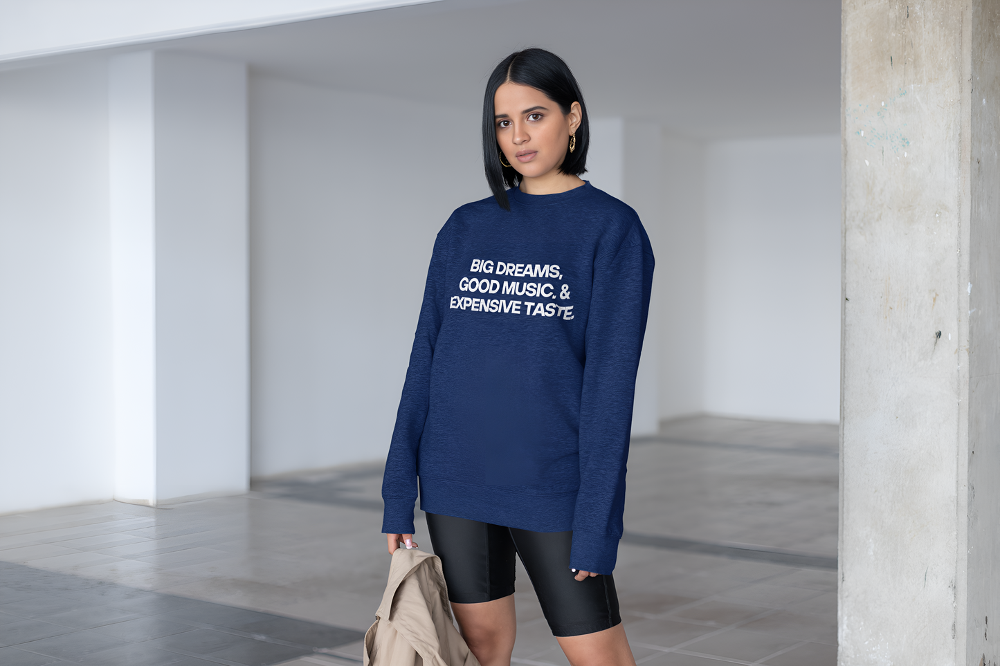 An Indian woman wearing a navy blue sweatshirt with "Big Dreams, Good Music, & Expensive Taste" written on it and shorts.