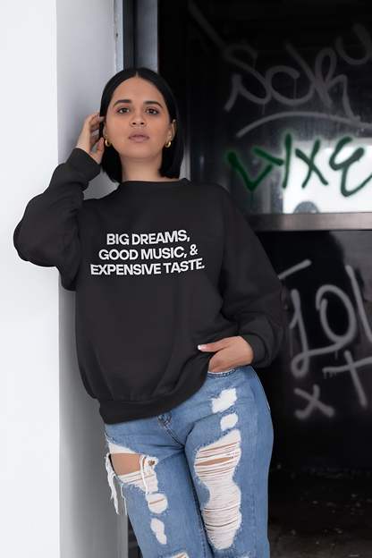 An Indian woman wearing a black sweatshirt with "Big Dreams, Good Music, & Expensive Taste" written on it standing in front of graffitti.