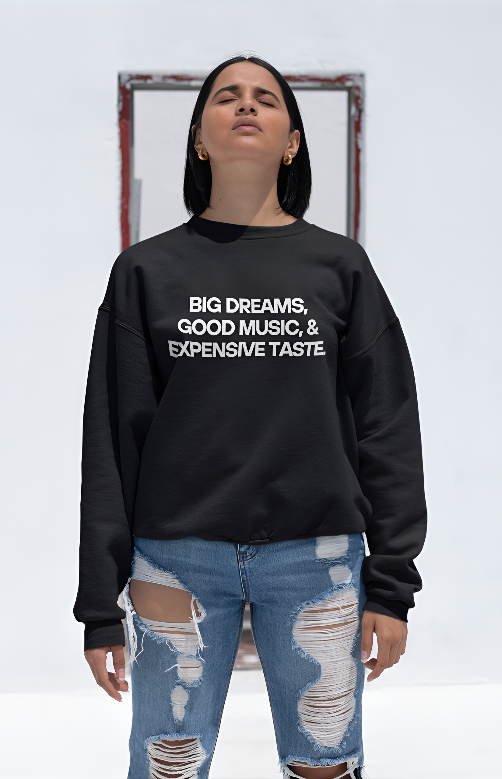 An Indian woman wearing a black sweatshirt with "Big Dreams, Good Music, & Expensive Taste" written on it looking up