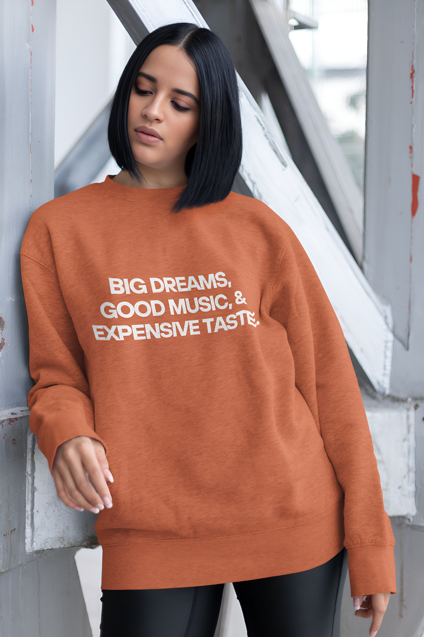 An Indian woman wearing a coral sweatshirt with "Big Dreams, Good Music, & Expensive Taste" written on it.